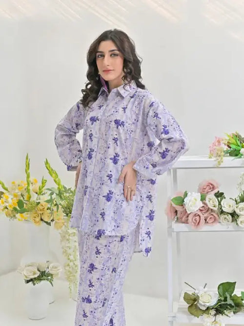 Woman wearing a matching lavender floral print shirt and trousers set, standing indoors with flowers in the background.
