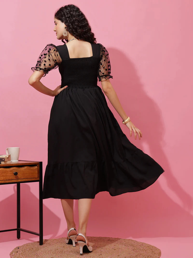 Woman in black midi dress with sheer polka dot sleeves, standing on a rug by a side table