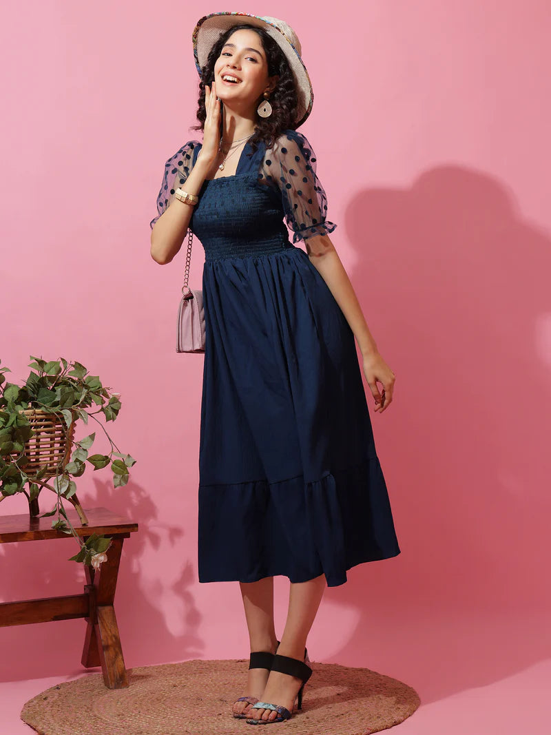 Woman in navy blue midi dress with sheer polka dot sleeves, straw hat, and heels against pink background