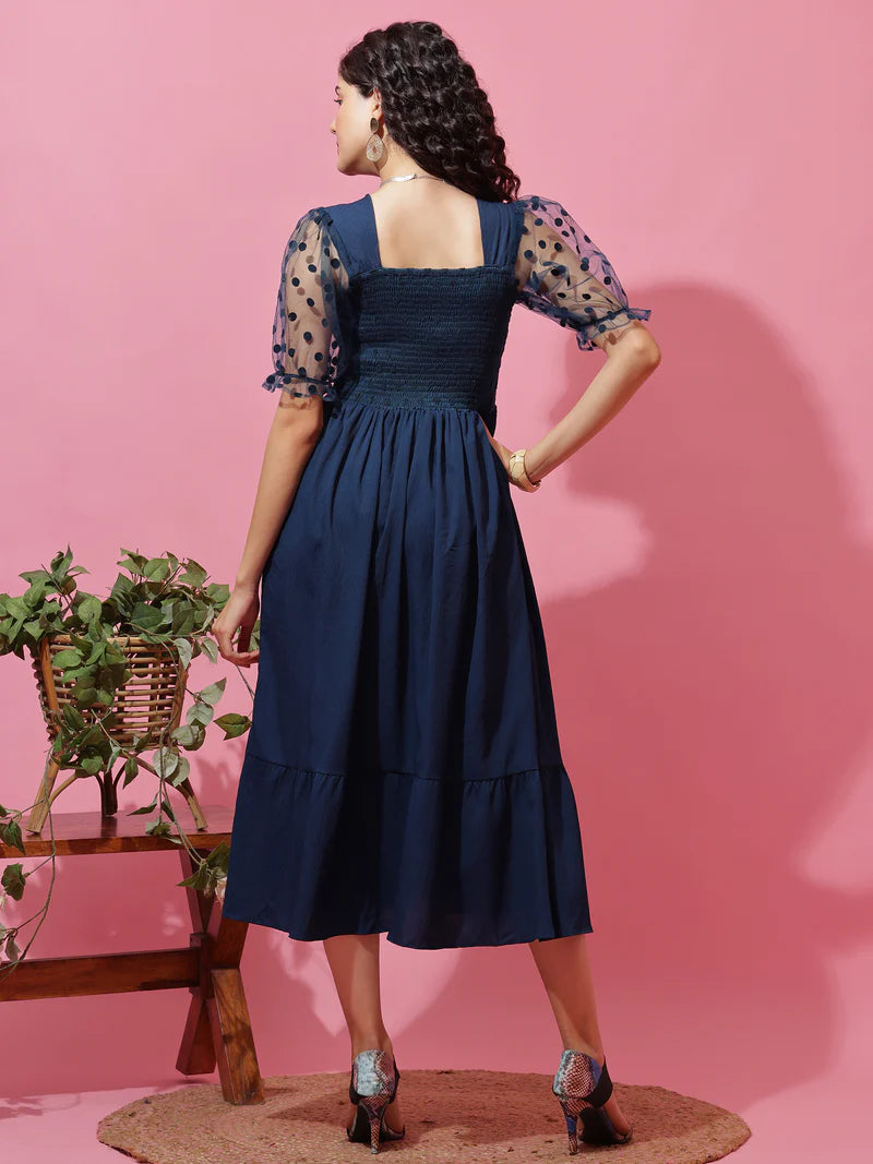Woman wearing navy blue midi dress with sheer polka dot sleeves, standing on pink background