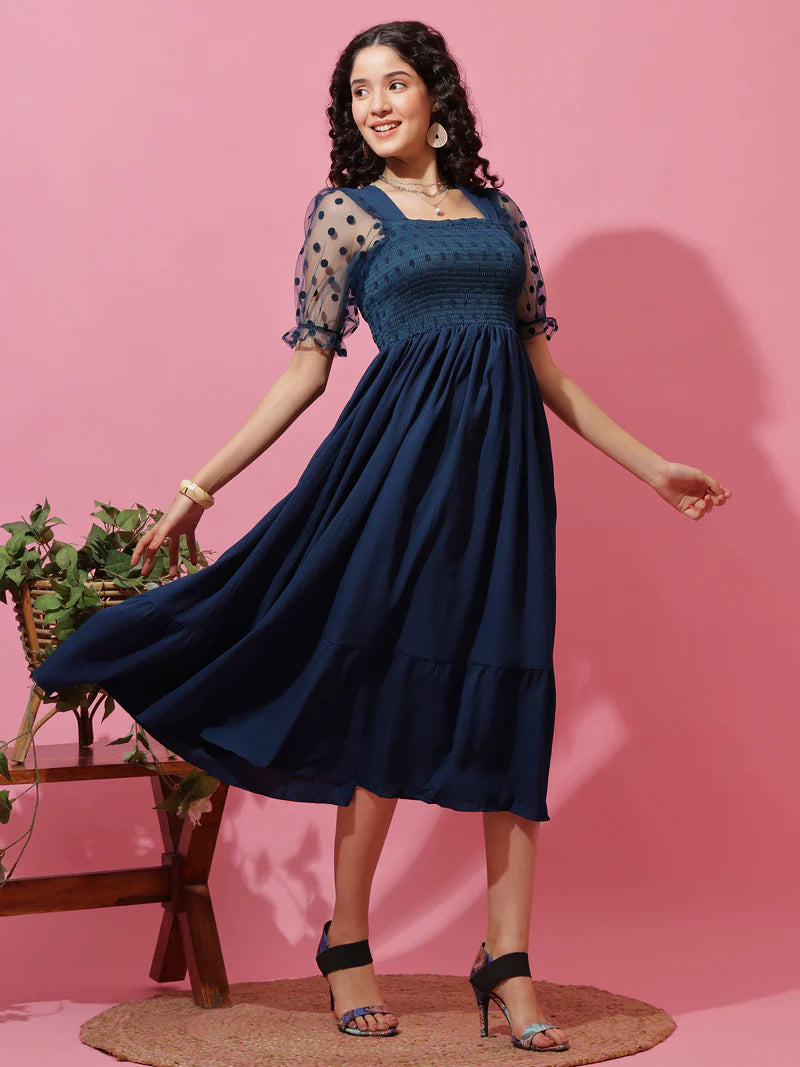 Woman wearing navy blue midi dress with polka dot sheer sleeves, posing against a pink background