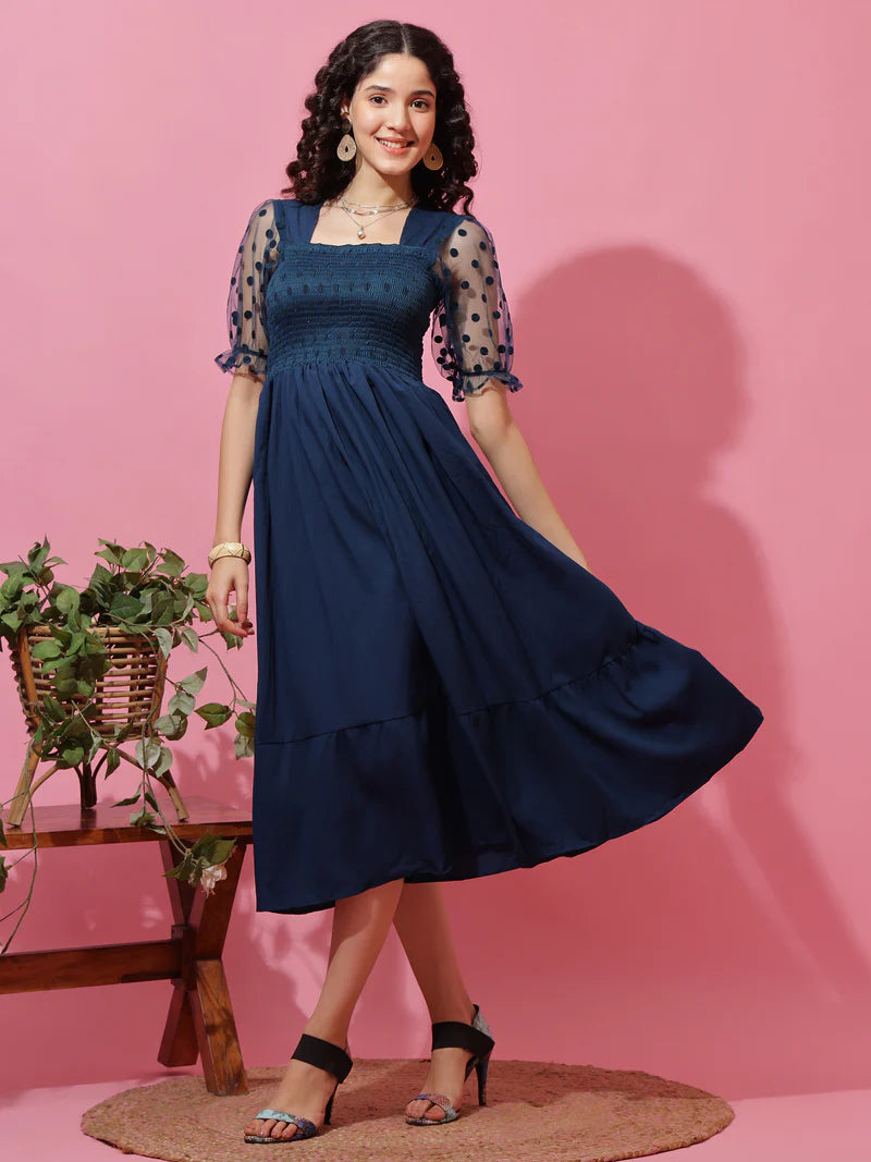 Woman wearing a navy blue midi dress with sheer polka dot sleeves, standing on a pink background.
