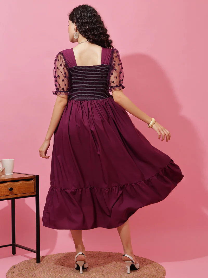 Woman in a purple midi dress with polka dot sheer sleeves, standing by a wooden side table.